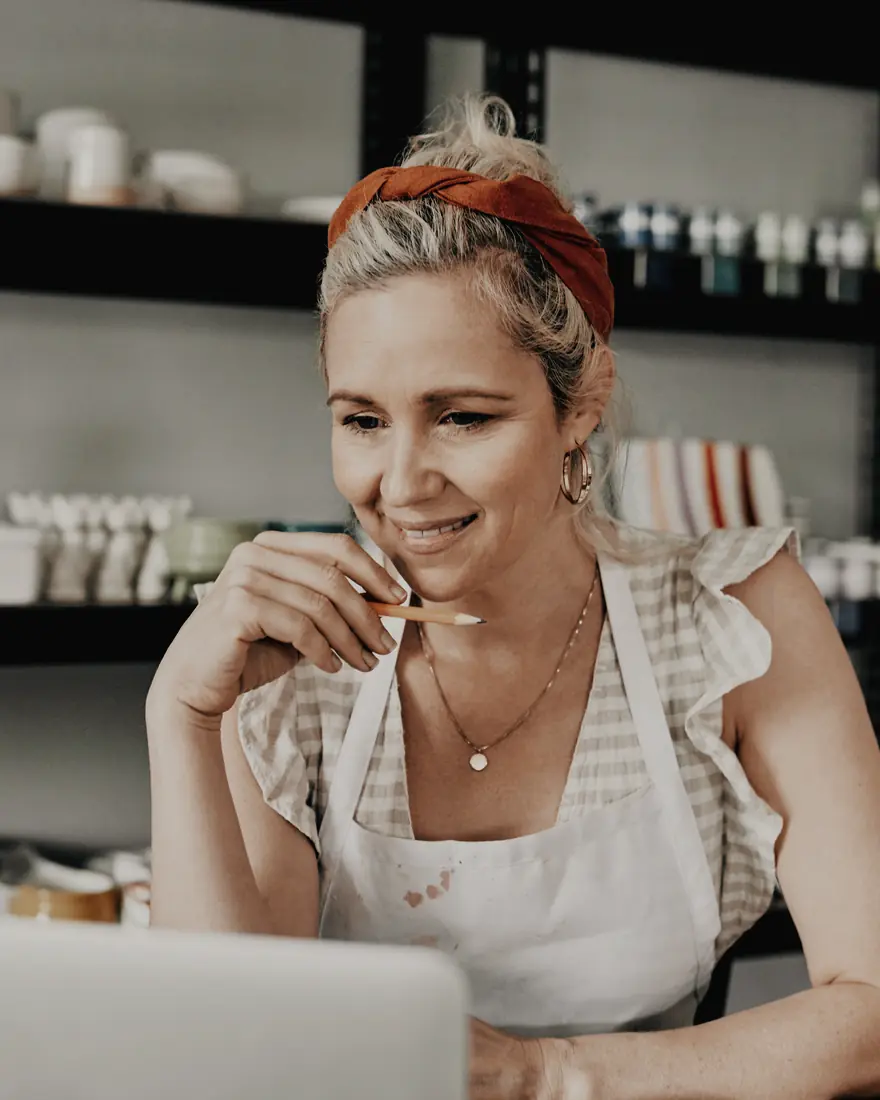 Kobieta w fartuchu siedzi przy laptopie w pracowni ceramicznej; za nią półki z kubkami i szkliwami.